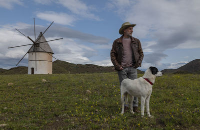 Adult man in cowboy hat and his dog standing on field  with traditional windmill in background