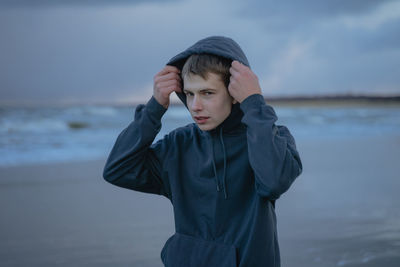 Handsome teenage boy in blue hoodie and hoodie standing outdoors against the background of the sea 