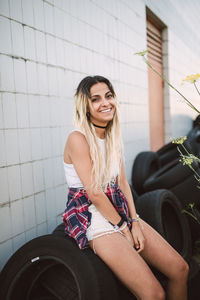 Portrait of young woman sitting on tire against tiled wall