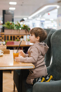 Four year old boy sits at a table in a cafe and looks at a glass of orange juice, holding a straw