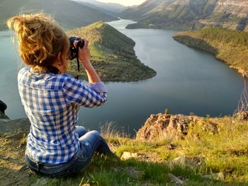 Rear view of mature woman photographing lake while sitting on mountain