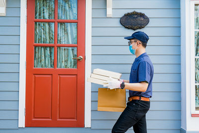 A male delivery man holds a box of goods and food for a customer in front of the house.