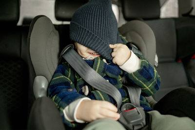 Happy boy covering face with knit hat in car safety seat