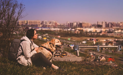 A girl sits hugging a labrador retriever on a mountain against the background of the city