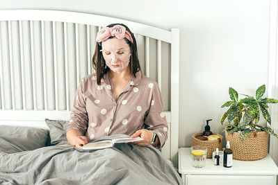 Woman with face mask reading book while while sitting on bed at home