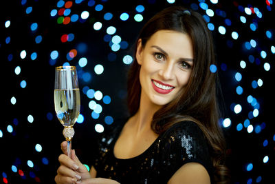 Portrait of beautiful young woman holding champagne flute against illuminated lights