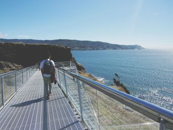 Rear view of man walking by sea on footbridge during sunny day