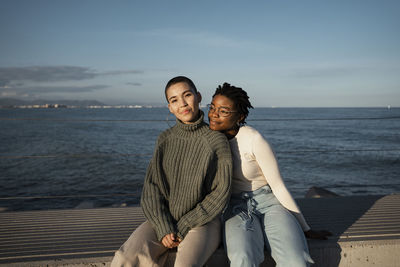 Woman leaning on friend shoulder while sitting on bench against sky