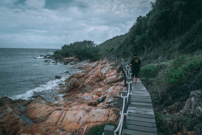 Rear view of woman walking on boardwalk at beach against cloudy sky