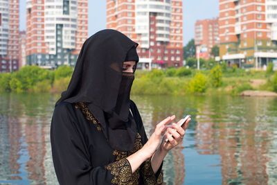 A muslim woman in national clothes with a phone  against the background of river in a megalopolis.