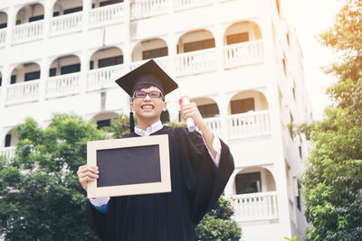 Smiling young man in graduation gown holding writing slate against building