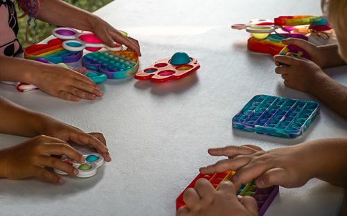 Popit and simple dimple toys among hands of playing children at white table.