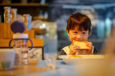 Little child, boy, sitting in the dining room eating bread, for breakfast on a sunny morning. 