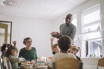 Male teacher serving food to children sitting at dining table during lunch break at kindergarten