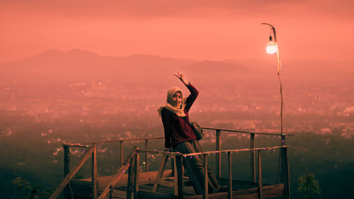 Woman sitting on rooftop by railing against sky during sunset