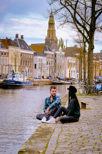 Man and woman sitting by canal against buildings and sky