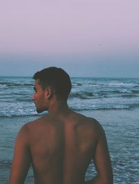 Rear view of shirtless young man standing at beach against sky