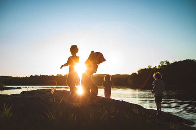 Children playing by lake against sky during sunset