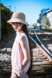 Side view of woman wearing hat standing against railroad tracks