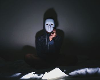 Portrait of young man wearing mask while sitting on bed in darkroom