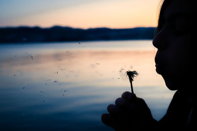 Close-up of silhouette hand holding dandelion against sky during sunset