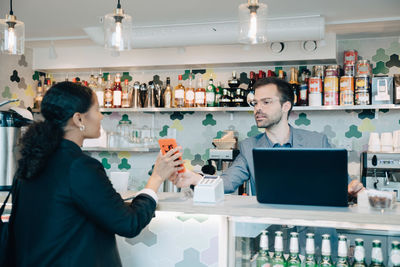 Businesswoman showing smart phone to owner at checkout counter in office cafe