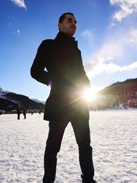 Low angle view of young man with hands in pockets standing on snow covered field against sky