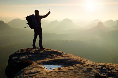 Tourist with backpack takes photos with smart phone on sharp rocky peak. dreamy foggy valley below