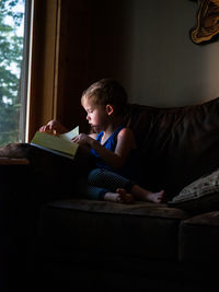 Boy reading book while sitting on sofa at home