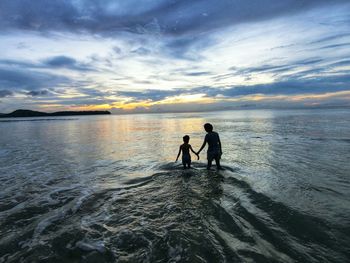 Mother and son wading in sea against sky during sunset