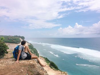 Side view of man sitting on rock at cliff by sea against sky