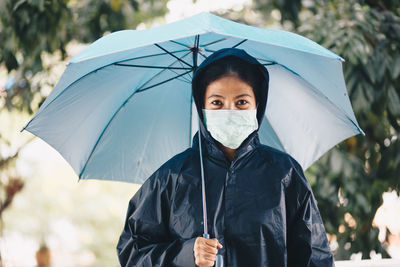 Portrait of woman wearing mask and raincoat holding umbrella whole standing outdoors