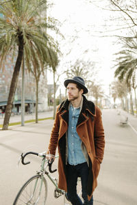 Hipster man wearing hat standing with bicycle in city while looking away