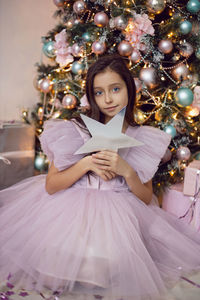 Girl child in a pink dress sits at the christmas tree and holds a star in her hands near her face