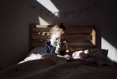Girl embracing doll while sitting on bed in darkroom at home