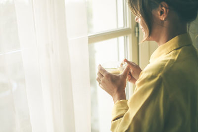 Young woman drinking a cup of tea looking outside the window girl enjoying free time at home. 