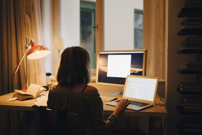 Rear view of girl using laptop and computer while sitting at illuminated desk