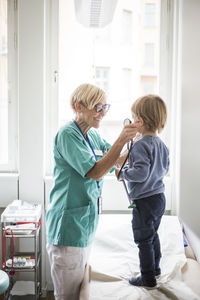 Smiling female doctor holding stethoscope to boy's ear while standing in hospital