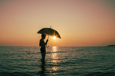 Young boy holding umbrella in sea during sunset