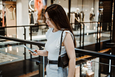 Young latina woman with shopping bags using her smart phone while standing in shopping center, mall