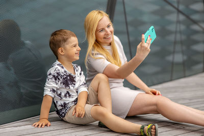 Happy blond woman and little boy sitting on terrace and making selfie on smartphone. mother and son