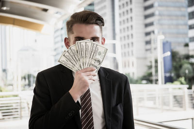 Businessman covering face with paper currency while standing in city