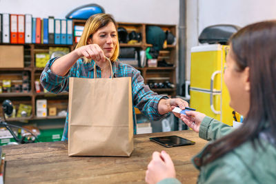 Smiling young woman shop assistant delivering paper bag with purchased items to female customer