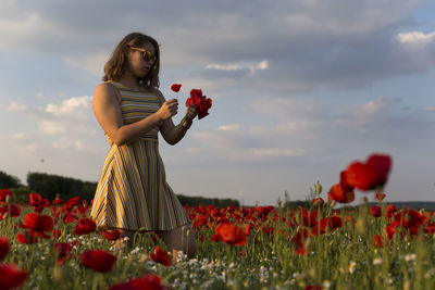 Woman standing by red poppy flowers on field against sky