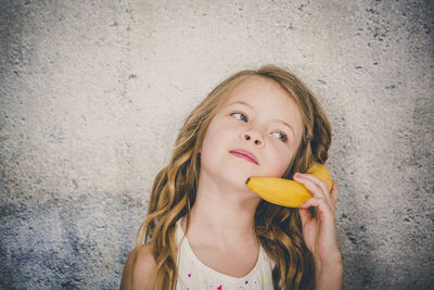 Close-up of cute girl using banana as phone while playing against textured wall