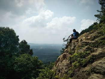 Side view of mid adult man sitting on rocky mountain against cloudy sky