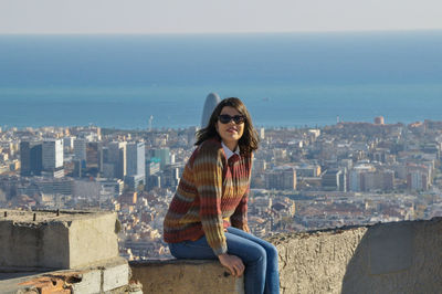 Young woman sitting on wall against cityscape and sea