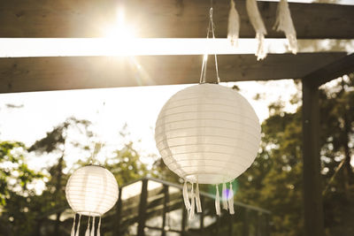 Low angle view of white lanterns hanging from pergola at patio on sunny day