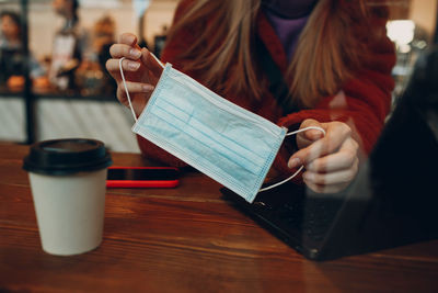 Midsection of woman holding mask while using laptop in cafe seen through window