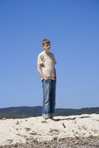 Full length of young man standing with hands in pockets on sandy beach against clear blue sky